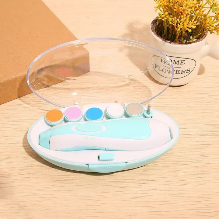 A mint green and white electric nail care set with five colorful attachments (orange, pink, blue, white, silver) in an oval case with a transparent lid, placed on a wooden surface beside a small potted plant labeled “Home Flowers.”
