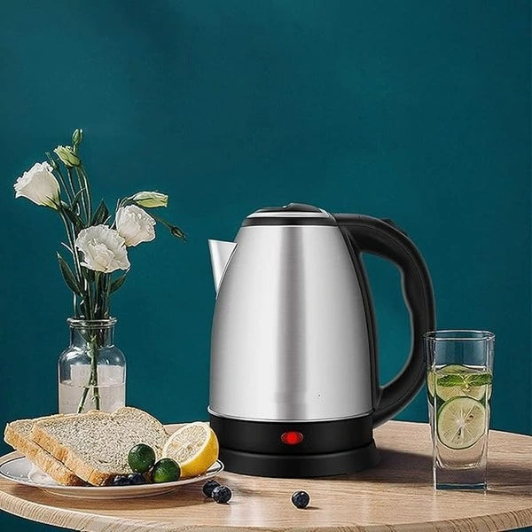 Silver electric kettle on a wooden table with a plate of toast, lemon, and berries, a glass of lemon water, and a vase of flowers against a teal background.